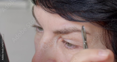 A woman meticulously performs eyebrow grooming with tweezers, emphasizing the importance of precision and skill in maintaining perfectly shaped eyebrows for an elegant look