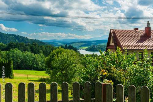 A wooden fence and red-roofed houses overlook a valley with a blue lake and historic castle in the distance. A tranquil landscape blending architecture, greenery, and Polish heritage.