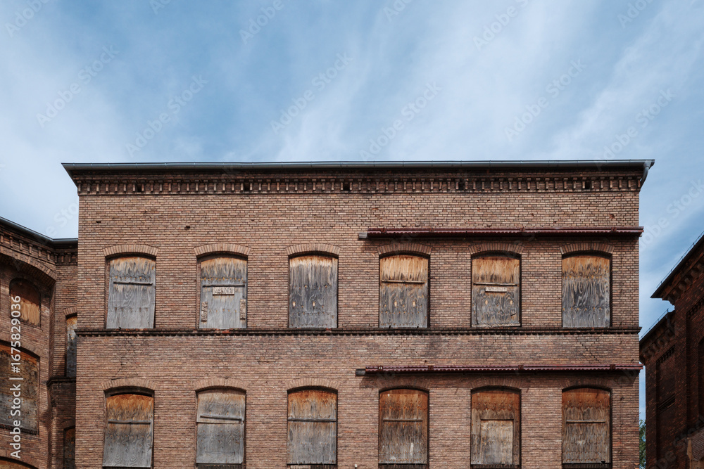 Fototapeta premium The weathered brick facade of an old, abandoned industrial factory or mill under a blue sky. All of the arched windows on the upper floors have been boarded up with old wood