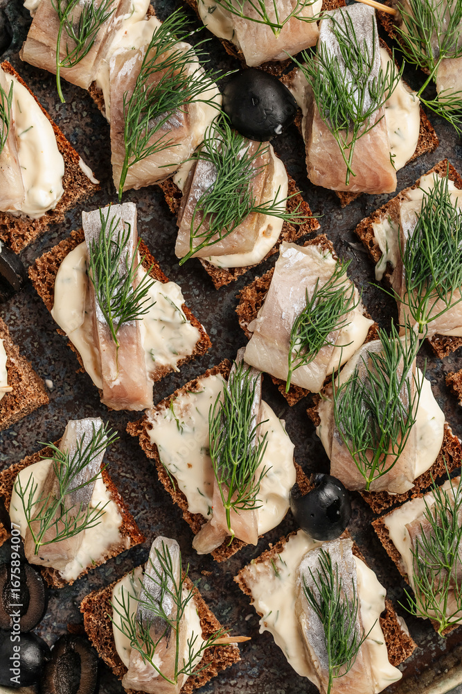Obraz premium Traditional sandwiches with black rye bread and salted herring, served with onion and herbs. Classic Eastern European appetizer on a rustic wooden table