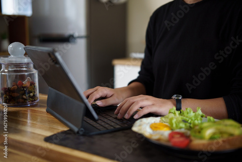 Wallpaper Mural Young woman working on digital tablet in the kitchen while having a healthy breakfast with eggs, salad, and avocado toast. Torontodigital.ca