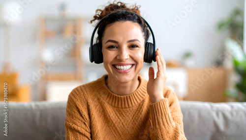 A cheerful woman wearing headphones smiles while looking directly at the camera, sitting on a sofa in a cozy home setting.