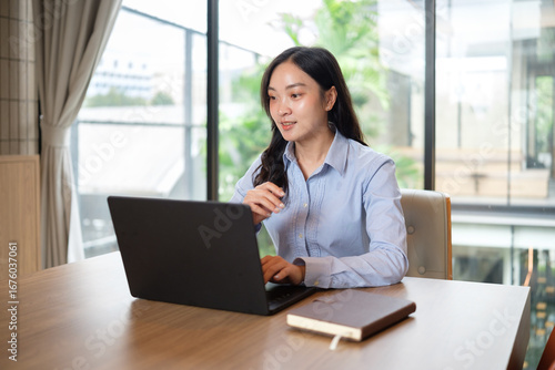 Businesswoman working on a laptop at wooden desk in a modern office with natural light.