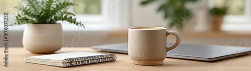 A ceramic mug, notebook, laptop, and potted plant on a wooden desk in a bright, cozy workspace.