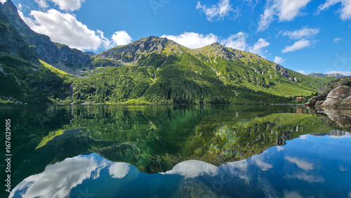 Fotografie Crystal clear mountain lake perfectly reflecting green slopes, forests, and bright clouds, creating breathtaking symmetrical wilderness scenery