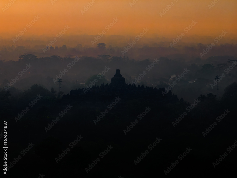 Fototapeta premium The iconic silhouette of Borobudur Temple emerges from a mystical morning mist, set against the warm, glowing colors of the dawn sky.