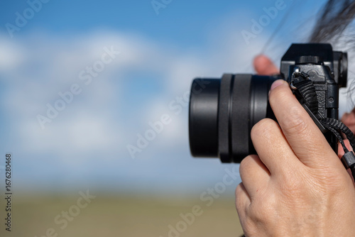objectif d'un appareil photo tenu en main par une photographe sur ciel bleu avec zone d'écriture
