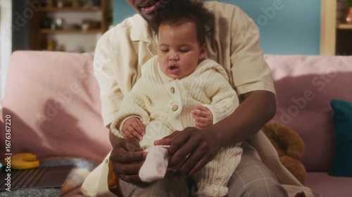 Close up shot of cute baby girl sitting in lap of smiling African American father caring for daughter and putting on pink socks