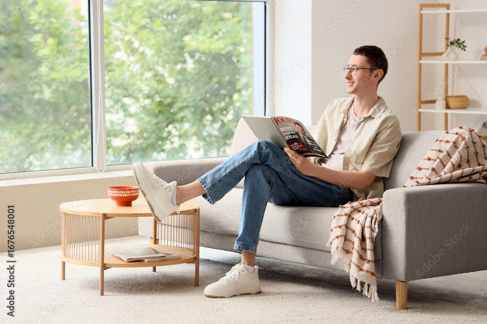 Obraz premium Young man in eyeglasses reading magazine on sofa at home