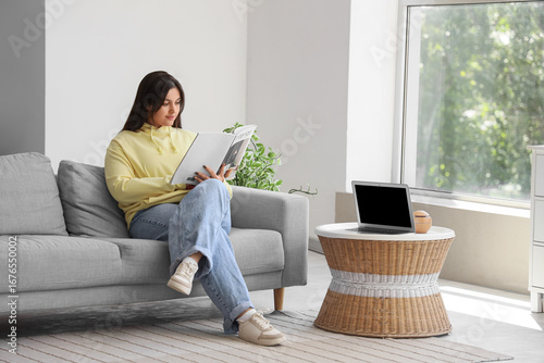 Beautiful young woman reading magazine on sofa at home