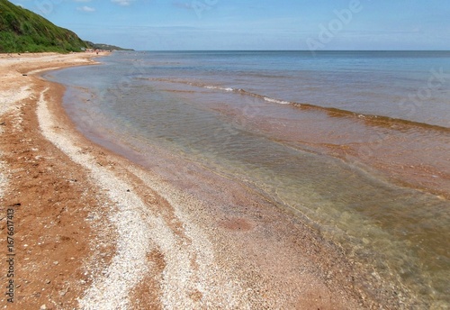Clear shallow water of a sandy sea beach with shell gravel. Green grassy hills and blue sky.