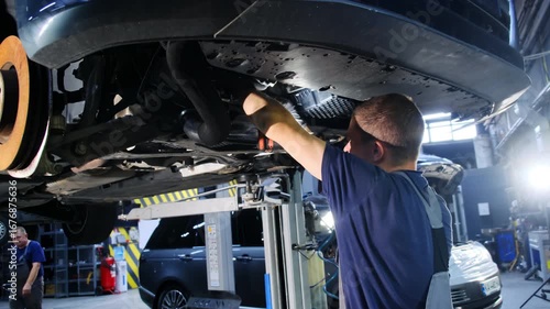 Man works under vehicle analyzing extent of malfunction. Mechanic and colleague repair car using special work llift to assess details