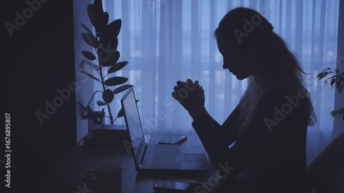 dark unrecognizable silhouette unhappy tired woman working on laptop sitting at desk in black room window light. Sad girl suffers from headache depression fatigue lack of money, Person looking for job