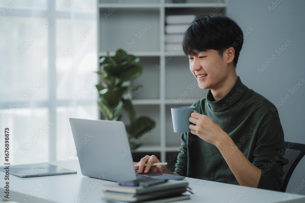 Obraz premium Asian freelancer working remotely using laptop and drinking coffee from mug, sitting at desk in home office