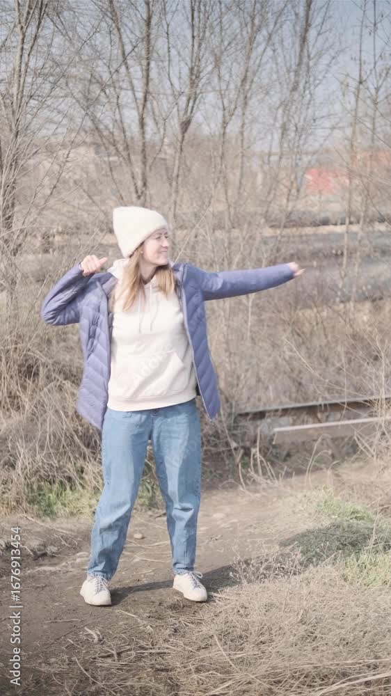Cheerful woman happily dancing outside near road, enjoying sunny day wearing warm jacket and hat Cheerful woman happily dancing outside near road, enjoying sunny day wearing warm jacket and hat