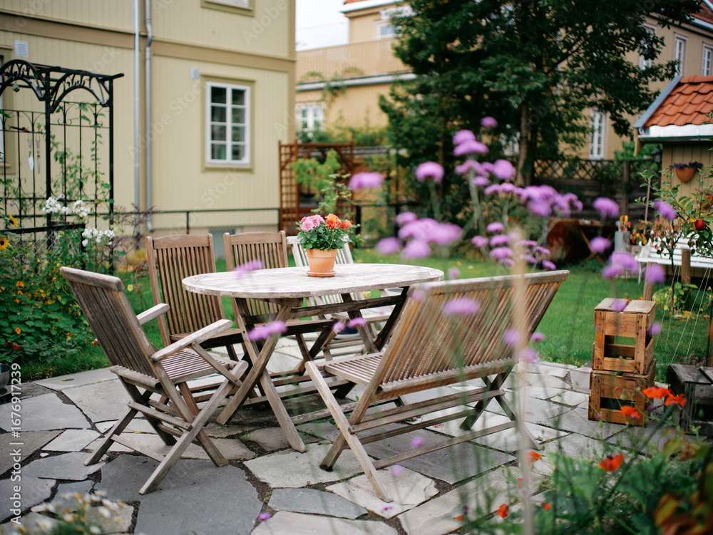 Fototapeta premium Charming Backyard Oasis with Wooden Patio Furniture Set Surrounded by Vibrant Blooming Flowers and Lush Greenery in a Cozy Outdoor Setting
