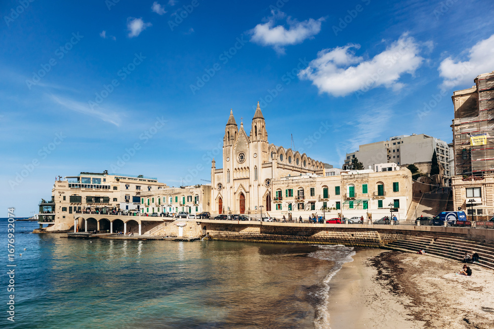 Obraz premium Balluta Bay, Malta – April 9, 2025: The neo-gothic Carmelite Church stands on the waterfront of Balluta Bay between St Julian's and Sliema, surrounded by traditional and modern buildings