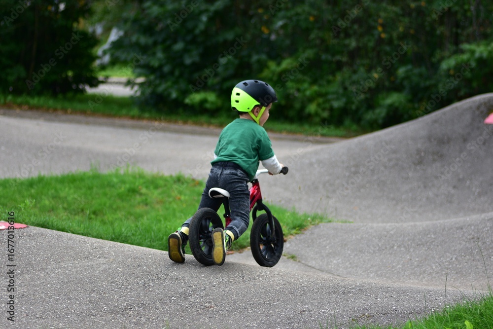 Obraz premium Toddler boy wearing helmet rides red balance bike on asphalt pump track. Active child learning balance and coordination while enjoying outdoor play in park.