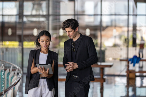 Young business officers in black jacket exchange ideas on business using tablet to provide data and information at a terrace outside of the building. Conversation of two coworkers in casual dress