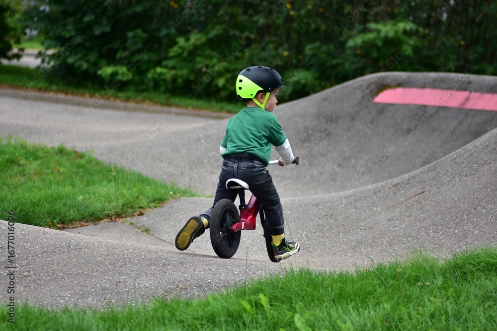Obraz premium Toddler boy wearing helmet rides red balance bike on asphalt pump track. Active child learning balance and coordination while enjoying outdoor play in park.