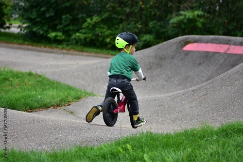 Toddler boy wearing helmet rides red balance bike on asphalt pump track. Active child learning balance and coordination while enjoying outdoor play in park.