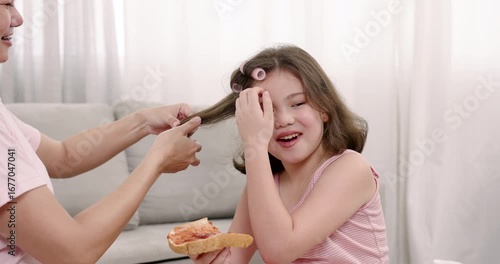 Asian adult female rolls hair of multiracial girl aged 8 who holds pizza calmly while sitting on sofa during beauty morning routine showing parenting care balance of grooming and comfort