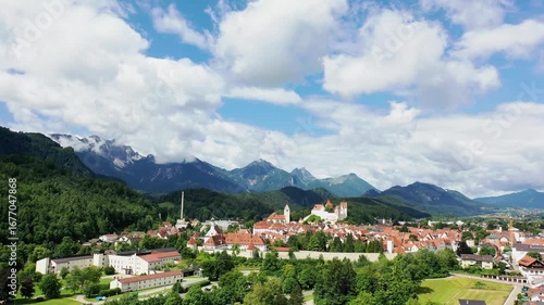 Das Foto zeigt die charmante Altstadt von Füssen mit ihren roten Dächern, liebevoll restaurierten Fassaden und dem markanten Turm der Kirche St. Mang. Im Hintergrund erhebt sich das beeindruckende Alp