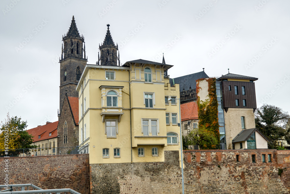 Naklejka premium Yellow building at Schleinufer with Magdeburg Cathedral in the background under a gray overcast sky.