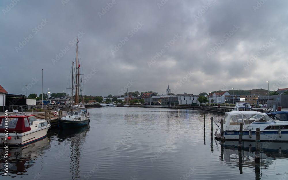 Fototapeta premium Boats and yachts docked in Thisted marina under dramatic clouds. View of the harbor and town with church spire, colorful houses, and reflections on the water in North Jutland, Denmark