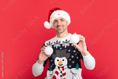 Santa with bauble, christmas ball. Handsome man in a winter sweater and Santa hat on studio red background.