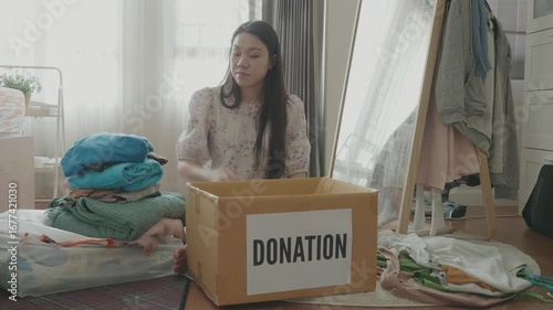 A young woman sorts through clothes and items in her living space, preparing a donation box. The scene highlights the importance of decluttering and giving back to the community through charitable don