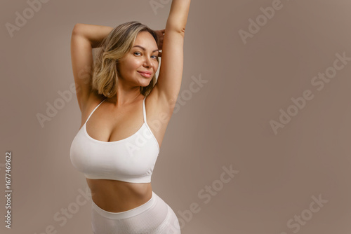 Young beautiful woman in white lingerie, smiling and touching her body, showing natural beauty, breast care, body positivity and healthy lifestyle on beige background.