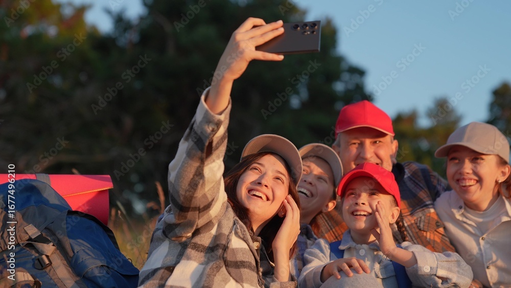 Fototapeta premium Happy travelers taking photos on smartphone at halt laughing hugging friend. Family trip with backpacks in nature. Team of travelers, rest stop, video bloggers. Team of tourists on vacation nature