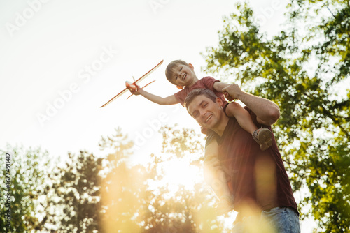 Fototapeta A father carries his excited son on his shoulders in a park, the boy holding a toy airplane and imagines flying as they enjoy a fun, carefree moment together