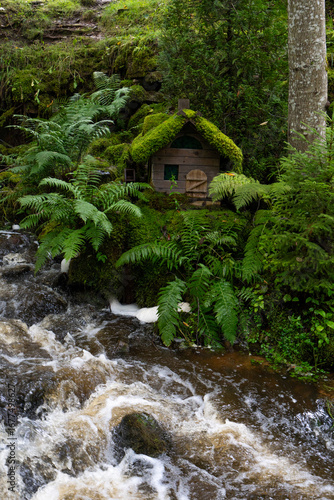 The image shows a small waterfall scene near the ruins of the Ieriķi Mill, where a stream flows through a lush green forest.