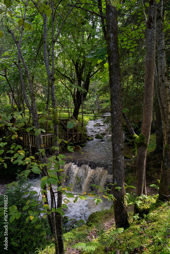 The image shows a small waterfall scene near the ruins of the Ieriķi Mill, where a stream flows through a lush green forest.