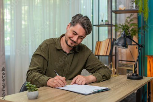 Billede på lærred Middle-aged man at home reviews documents smiles satisfied signs papers approving content