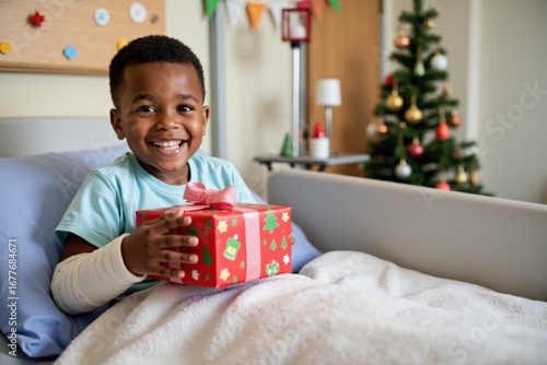 A black boy with a cast on his arm sits in a hospital bed, happily opening a Christmas present. The room has colorful decorations and a small Christmas tree