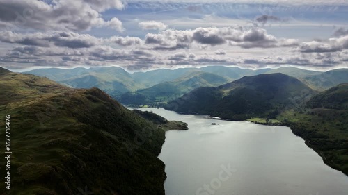aerial panning left drone shot over lake inside mountain valley with hills covered in trees and grass in England Lake District in summer with blue skies