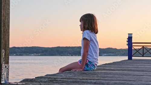 Cute Caucasian Girl Kid Sitting Alone on Wooden Pier at Serene Lake Yacht Harbor on Golden Hour Sunset Afternoon