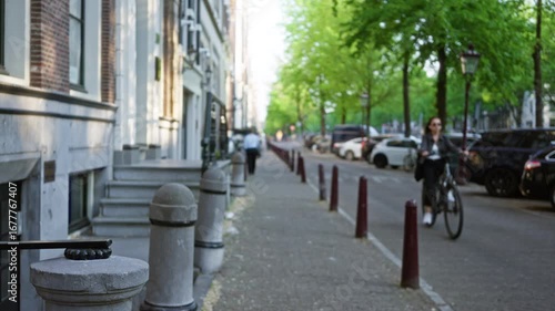 City street in amsterdam with a blurred man walking and woman cycling under green trees with cars parked along the sidewalk.