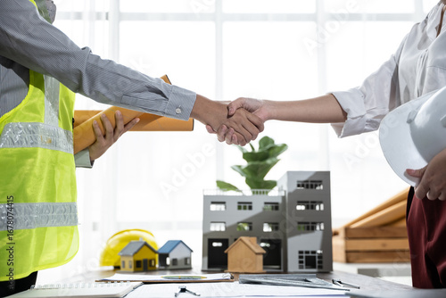 Two construction professionals shake hands over architectural models and paperwork, sealing a building project agreement in a bright office environment.