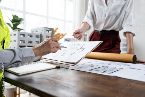 Architects review floor plans at a desk with scale models, blueprints, and tools in a sunlit office, discussing construction details and project planning.