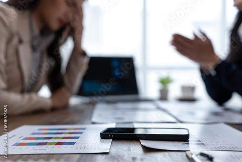 Smartphone and financial charts on a conference table as colleagues debate in the background, suggesting a tense business meeting and data review.