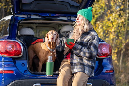 Woman pet owner drinking tea sitting in car trunk with dog enjoying nature in forest on weekend. Autumn outdoors activity in cold weather, vacation travel tourism road trip with magyar vizsla dog.