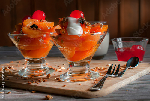Apricots with granola and yogurt in a glass cup with a cherry on top on a wooden cutting board, and a wooden wall out of focus in the background, side view