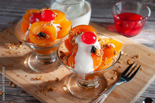 Apricot dessert in a glass cup with granola, yogurt, and a cherry on top on a wooden cutting board with a fork angle side view.