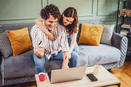 Obraz na plátně A smiling couple uses a laptop to pay for online shopping, enjoying a fun and relaxed moment together in the living room