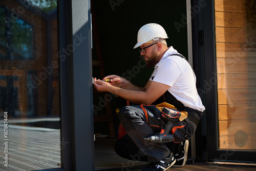 Construction worker installing door in a new house construction site. Man in working attire install front door. Builder in uniform with screwdriver adjusting front glass door in new beautiful house.