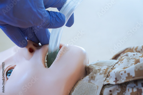 Medical training mannequin in camouflage uniform during insertion of an i-gel supraglottic airway device. The procedure illustrates airway management placement as part of a military medical simulation